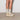 En model som bär beiga strumpor och ett par MoEa GEN 2 Corn Full Sand veganska sneakers i en kombination av vitt och beige. Skorna är tillverkade av majsskinn. De har en låg profil med snörning framtill och beige snören. Den vita färgen dominerar, med beige detaljer runt tån, hälen och sidopanelerna. På sidan av skon finns 'MoEa'-logotyp. Sulan är beige, tjock och räfflad, gjord av återvunnet gummi. Skorna har också perforerade detaljer på sidorna för ökad ventilation.